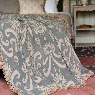 blue linen throw with an Usbek style print and a tassel trim, draped over the edge of a bed. 