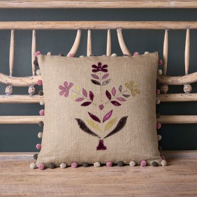 Square linen cushion appliqued with a floral design in shades of charcoal and purple and a pom pom trim. 